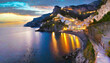 A rocky coastal scene during a winter evening in the Amalfi Coast, Italy, with the twinkling lights of seaside villages and the Tyrrhenian Sea reflecting the fading sunlight.