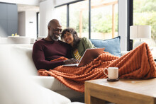 Happy diverse mature couple relaxing on couch with blanket using laptop in sunny living room
