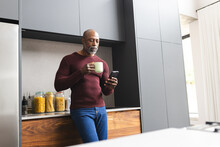 Focused mature african american man having coffee using smartphone standing in sunny kitchen