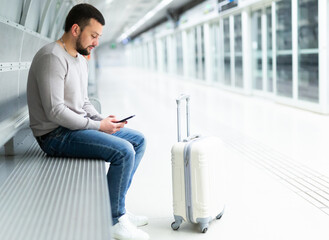 Young bearded guy traveler waiting for train at metro station sitting on bench with suitcase, chatting on social networks on his phone..