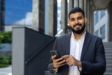 Wall Mural - Close-up photo of a smiling young Arab man standing on the street near the office, holding a phone and smiling at the camera