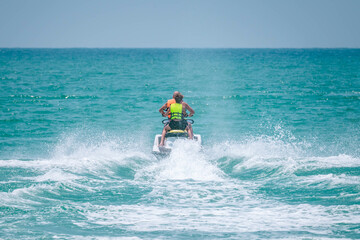 Two persons wearing safety life jackets drive jetski from shore to open sea, splashing water. Tropical beach resort. Close up photo