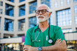 © Jelena Stanojkovic - Portrait of a successful mature doctor with a beard and in a medical uniform, outside a modern clinic, a hospital director with crossed arms smiling and looking away.