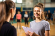 © MVProductions - Portrait of physical education female teacher in a gym hall smiling and holding a clipboard with pupils in the background