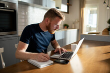 Bearded man using laptop taking notes at home