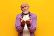 offended bald grandfather in purple shirt begs on yellow isolated background, sad old man pensioner holds empty hands
