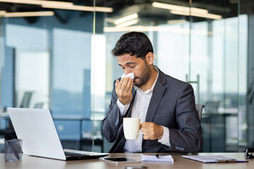 Young Hindu businessman sneezes and has a runny nose while working in the office at the computer, young man has a cold, drinks medicine, feels bad.