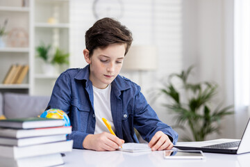 Concentrated student boy sitting at the table in front of the laptop at home, writing something in a notebook, doing homework, distance learning.