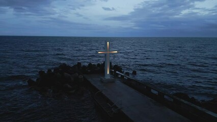 Wall Mural - A large Christian cross stands at the edge of a pier against a dramatic sky and sea, seen from above