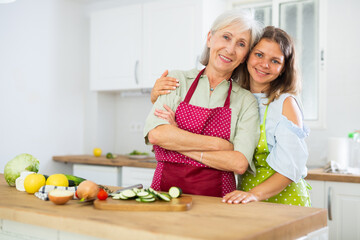 Wall Mural - Positive family, senior woman and her daughter in aprons, standing in kitchen in apartment. Preparing food at home.