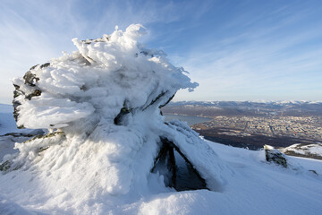 Wall Mural - View of snow-covered and icy rocks on a snow-covered mountain slope. In the distance is a large city located on the coast of a sea bay. Beautiful northern nature. Magadan, Magadan region, Russia.