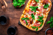 Rectangular roman pizza with smoked ham, mozzarella cheese, sun dried tomatoes, tomato sauce and green basil leaves on rustic wooden table background, top view