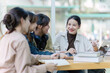 © itchaznong - Group of young Asian college students sitting on a bench in a campus relaxation area, talking, sharing ideas, doing homework or tutoring for the exam together