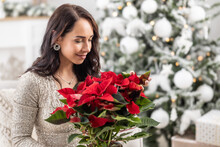 Woman Smiles And Smells A Poinsette In A Flower Pot Next To A Christmas Tree