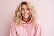 Young beautiful blonde girl with curly hair in pink hoodie laughs fervently on pink background. Human emotions, joy, laughter, happiness. Modern student