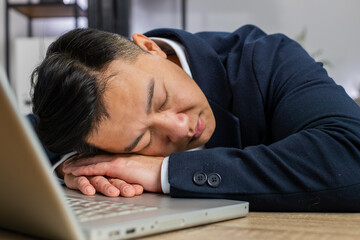 Bored sleepy Asian businessman worker working on laptop computer, leaning on hand falling asleep at office desk. Exhausted tired freelancer workaholic middle-aged man. Employment, occupation, workless