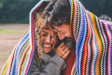 Intimate funny and happy moments with adult couple in outdoor leisure activity covered with a knit colorful cover at the park. Man and woman couple in love and relationship at the park together