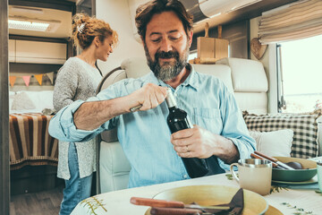 Adult traveler couple enjoy lunch time inside a camper van. Nomadic alternative lifestyle. Travel people in summer holiday vacation ready to eat. Man opening wine and woman cooking