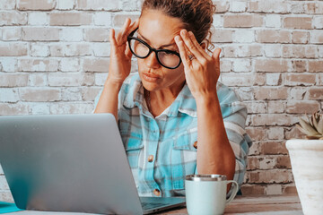 Wall Mural - Woman working at the desk on laptop with headache. Concentrated and worried female businesswoman in front of a computer. Home work place with wall bricks in background. Online job and security concept