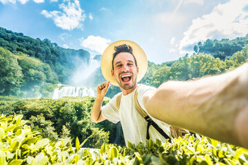 Wall Mural - Happy man with hat and backpack taking selfie portrait walking in the forest - Young traveler laughing at camera into the wild - Travel influencer recording social media video content