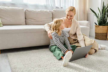 happy child with prosthetic leg using laptop and sitting on carpet with mother in living room