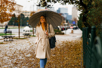 Wall Mural - Young woman with umbrella walking down a city street covered with leaves on a autumn rainy day