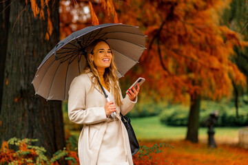 Wall Mural - Young smiling woman with white umbrella on a rainy autumn day looking at her phone on a stroll in park