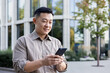 © Tetiana - Close-up photo of a smiling young Asian man, an office worker standing outside and using the phone