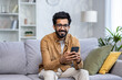 © Liubomir - Portrait of a young man at home, Indian man sitting on the sofa in the living room of the house, smiling, looking at the camera, holding a phone, using an application on a smartphone, typing a message