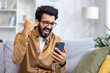 © Liubomir - Portrait of winner at home, happy happy man reading winning online notification, holding phone, holding hand up triumph gesture, satisfied with achievement results, sitting on sofa in living room.