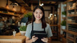 © MP Studio - A smiling woman, small business owner, holding a tablet and wearing an apron, standing in a well-lit and organized cafe environment