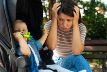 Tired Young Mother Sits On A Bench Upset Next To A Baby Stroller