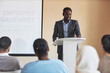© Seventyfour - Young African American male speaker in formalwear standing by podium and looking at audience while explaining points of his report