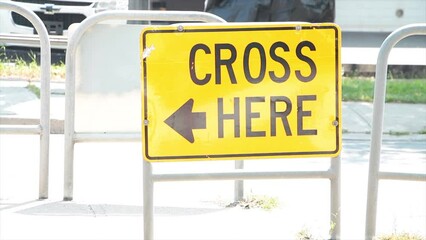 Sticker - cross here with arrow writing caption text street road sign on island in middle of road in black on yellow with cars vehicles traffic passing behind and in front