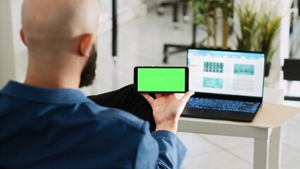 Poster - Worker looking at greenscreen display, preparing to do research for small business plan. Young adult holding smartphone with isolated copyspace mockup, open floor office space.