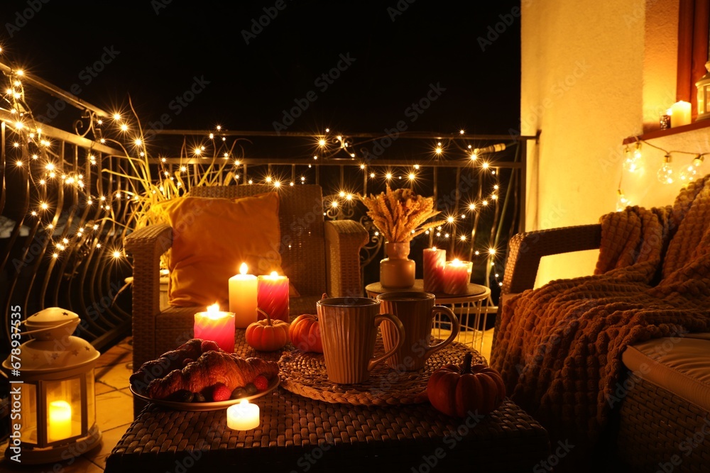 Rattan furniture, cups, fairy lights, burning candles and other autumn decor on outdoor terrace at night