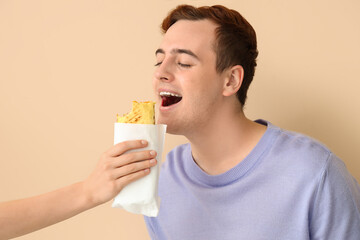 Wall Mural - Young man eating tasty sandwich wrap on beige background, closeup