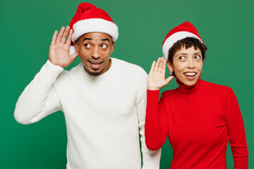 Wall Mural - Merry young couple friends man woman wear red casual clothes Santa hat posing try to hear you overhear listen intently isolated on plain green background Happy New Year 2024 Christmas holiday concept