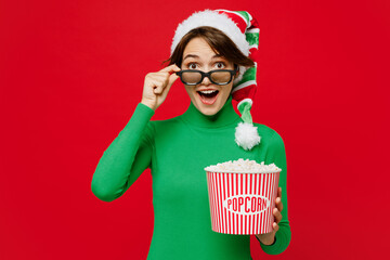 Wall Mural - Young surprised woman lower 3d glasses wear green turtleneck Santa hat watch movie film hold popcorn bucket in cinema isolated on plain red background. Happy New Year 2024 Christmas holiday concept.