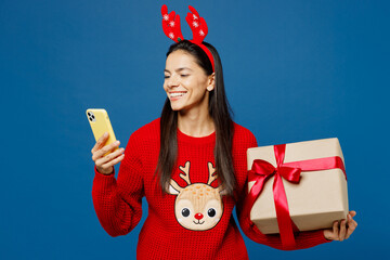 Wall Mural - Merry young Latin woman in red Christmas sweater decorative deer horns on head posing hold present box with gift ribbon bow mobile cell phone isolated on plain blue background Happy New Year concept