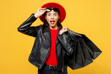 Young surprised shocked happy woman wear casual clothes red hat take off sunglasses hold shopping paper package bags look camera isolated on plain yellow background. Black Friday sale buy day concept.