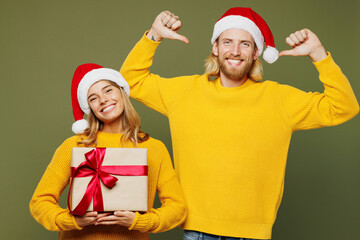 Merry young couple two friends man woman wear sweater Santa hat posing hold present box with gift ribbon bow point himself isolated on plain green background. Happy New Year Christmas holiday concept.