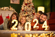 Parents and children lying by the Christmas tree holding numbers 2024 for New Year