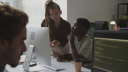 Medium shot of multiracial software experts looking at computer and having discussion of new program or app and programmer writing code in blurred foreground in modern office