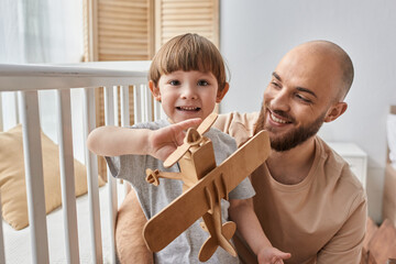 happy adorable boy playing with wooden plane with his father and smiling at camera, family