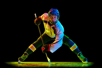 Concentrated young woman, hockey player un uniform and helmet, standing with stick against black studio background in neon light. Concept of professional sport, competition, game, action, hobby