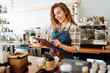 © maxbelchenko - Portrait of a friendly female barista holding a tablet computer and smiling. A young coffee shop owner standing behind the bar using a digital tablet. Takeaway concept, technology.