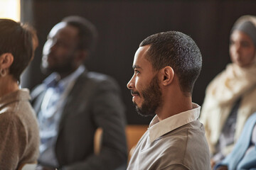 Side view of young bearded multiethnic businessman listening to report of speaker while sitting among intercultural audience at conference