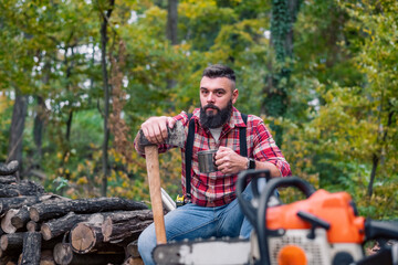 Wall Mural - Caucasian man, a lumberjack, takes a moment to rest on a pile of firewood, pouring himself a cup of coffee from a thermos, his tools laid out in front of him.