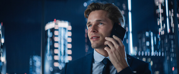 Poster - Close Up Portrait of Handsome Caucasian Businessman in Stylish Suit Standing Next to Window in Big City Office with Skyscrapers Late At Night. Male CEO Smiling, Talking On Smartphone.